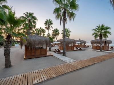 Strandbereich mit Liegen, kleinen Cabanas und Palmen bei Sonnenuntergang.