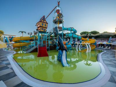 Aire de jeux aquatique colorée avec toboggans et seaux, entourée de chaises longues beiges au bord de la piscine.