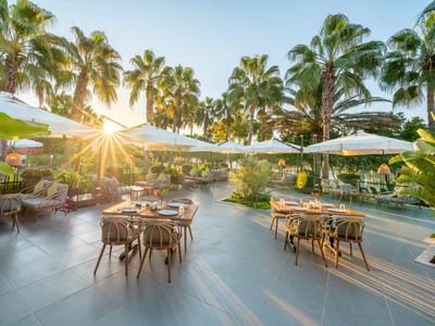 Außenbereich eines Hotels mit Tischen unter Sonnenschirmen und Palmen bei Sonnenuntergang.