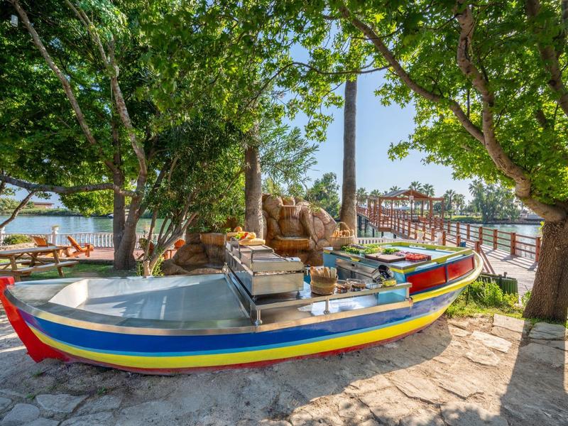 Bateau coloré posé sur une surface pavée sous des arbres avec vue sur l'eau et le port.