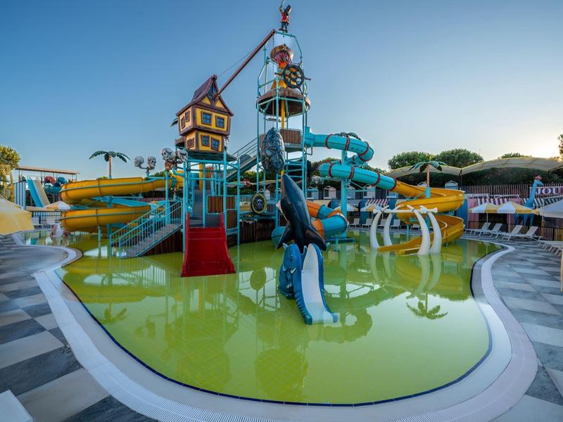 Aire de jeux aquatique colorée avec toboggans et seaux, entourée de chaises longues beiges au bord de la piscine.