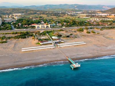 Langer Steg führt vom Strand ins ruhige blaue Meer, dahinter Sand und grüne Vegetation.