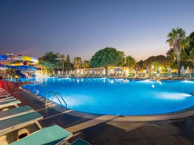 Großer Pool mit Wasserbeleuchtung, Liegestühlen und Palmen bei Dämmerung in einem Resort.