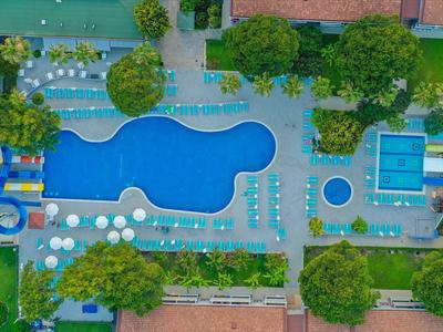 Luftaufnahme eines Hotelpools mit Liegestühlen und angrenzenden Gebäuden, umgeben von Bäumen.