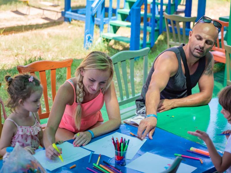 Vier Personen sitzen an einem blauen Tisch draußen und malen mit bunten Stiften auf Papier.