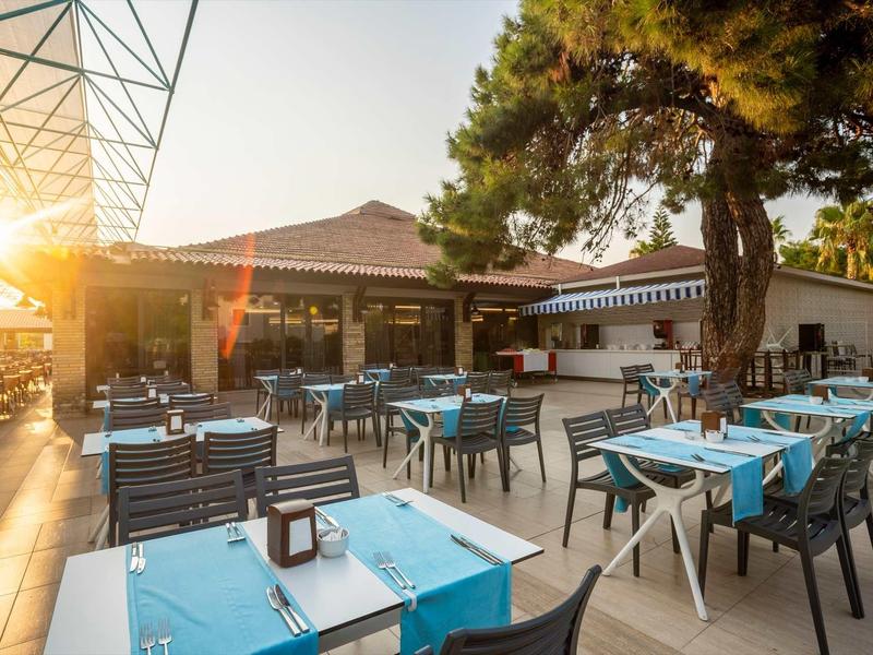 Freiluftrestaurant mit blauen Tischdecken, schwarzen Stühlen und großer Tanne bei Sonnenuntergang.