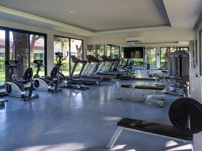 Heller Fitnessraum mit großen Fenstern, verschiedenen Trainingsgeräten und blauem Boden.