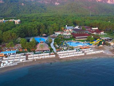 Luftaufnahme eines Resorts mit Strand, Pools und Wasserrutschen am Fuße eines bewaldeten Hügels.