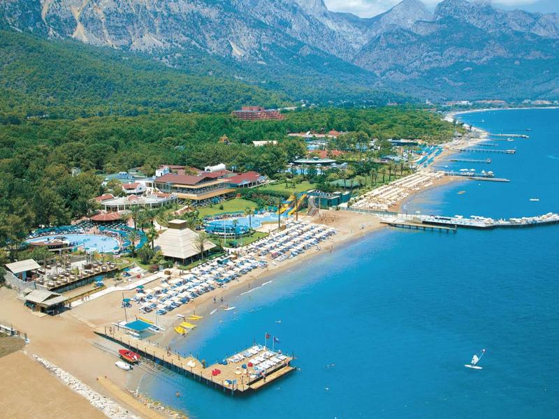 Vue aérienne d'un hôtel sur la plage avec des chaises longues, des jetées et des montagnes en arrière-plan.
