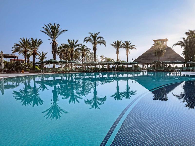 Grand piscine d'hôtel avec palmiers et parasols sous un ciel clair.