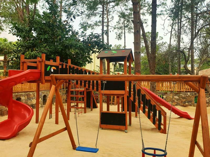 Aire de jeux pour enfants avec toboggan, balançoires et structure d'escalade en zone boisée.