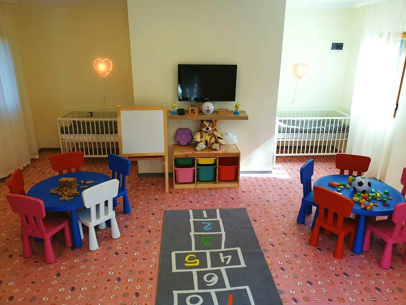 Salle de jeux lumineuse avec petites tables, chaises colorées et deux lits bébé contre le mur.