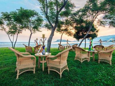 Wetterfeste Korbstühle mit Tischen stehen auf grünem Rasen unter Bäumen am Meer bei Sonnenuntergang.