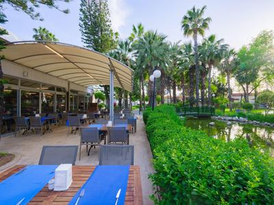Gartenrestaurant neben Teich mit grünen Büschen und Palmen, blauer Himmel, sonnig.