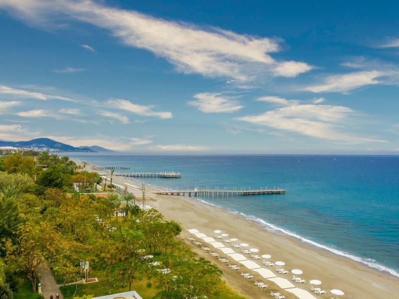 Strand mit Liegestühlen, grüner Vegetation und einem Steg bei klarem Wetter am Meer.
