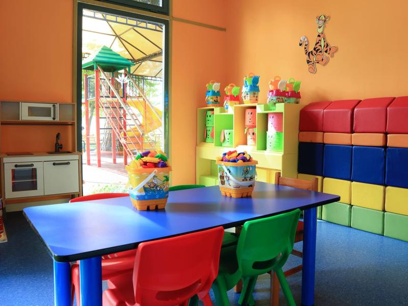 Bunter Kinderspielraum mit Tisch, Stühlen und Regalen voller Spielzeug neben einem Ausgang.