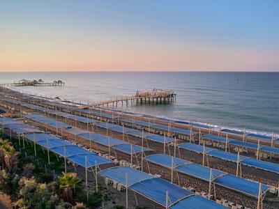Langer Steg ragt ins ruhige Meer, davor viele Reihen von Sonnendächern am Strand bei Sonnenuntergang.