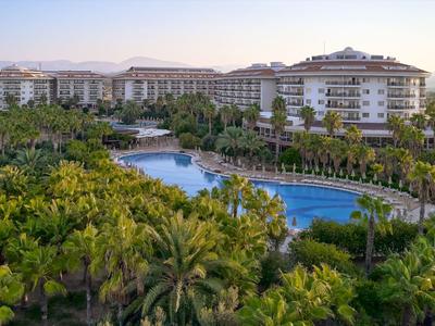 Große Hotelanlage mit Pool, umgeben von Palmen und Bergen im Hintergrund bei Sonnenuntergang.