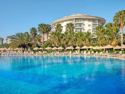 Großer Pool mit Liegestühlen und Palmen vor einem modernen Hotelgebäude bei blauem Himmel.