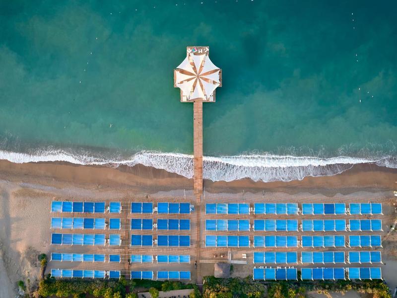 Übersicht über einen Strand mit blauen Liegen, einem Steg mit Sonnenschirm und türkisfarbenem Meer.
