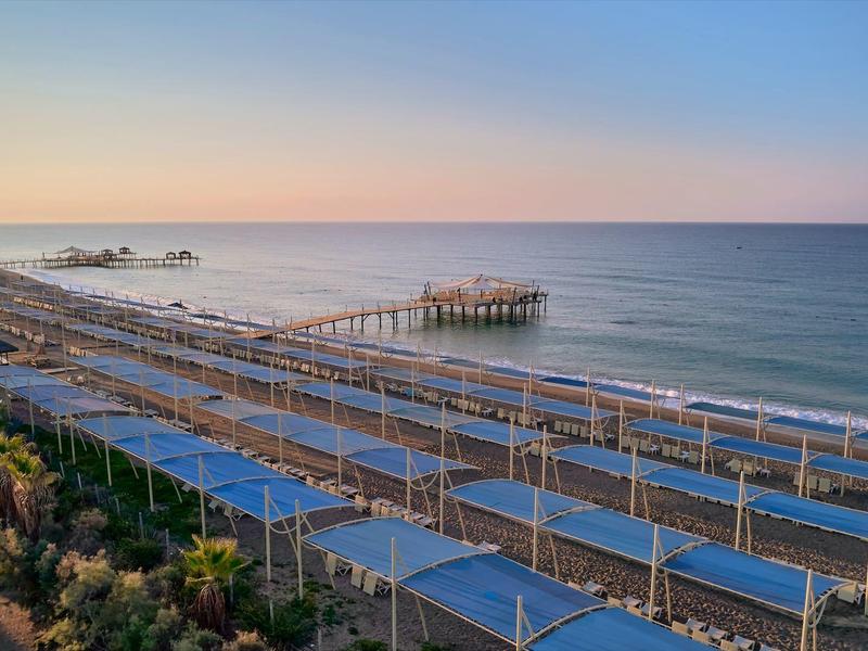 Langer Steg ragt ins ruhige Meer, davor viele Reihen von Sonnendächern am Strand bei Sonnenuntergang.
