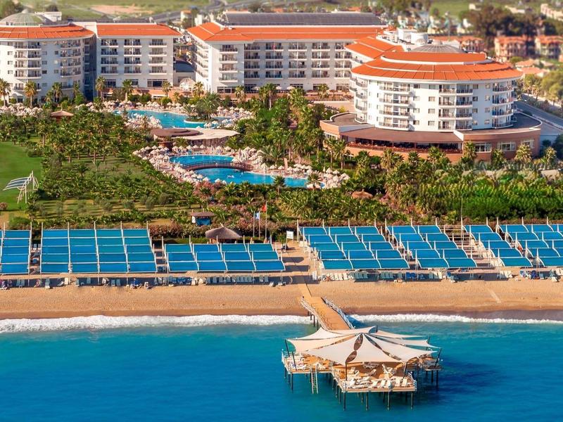 Großes Hotel mit roten Dächern, vielen Pools, blauen Liegestühlen am Strand und Steg ins blaue Meer.