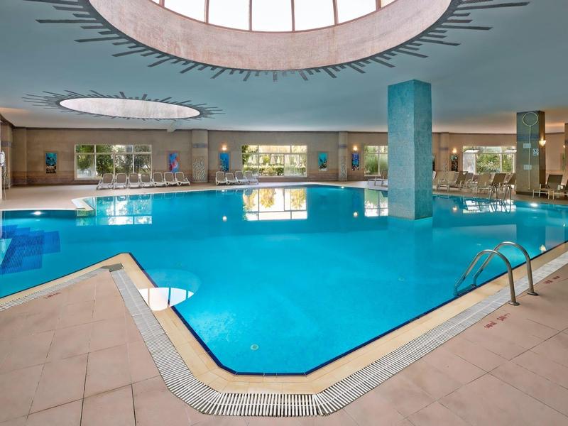 Großes Hallenbad mit blauem Wasser und runder Deckenbeleuchtung, umgeben von beige Fliesen.