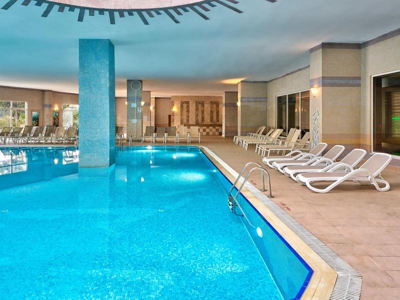 Innen-Schwimmbad mit blauem Wasser, weißen Liegestühlen und Fliesenboden.