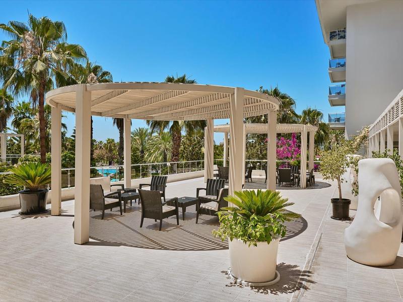 Moderner Außenbereich mit Pergola, Sitzmöbeln, Palmen und großen Pflanzenvasen bei blauem Himmel.