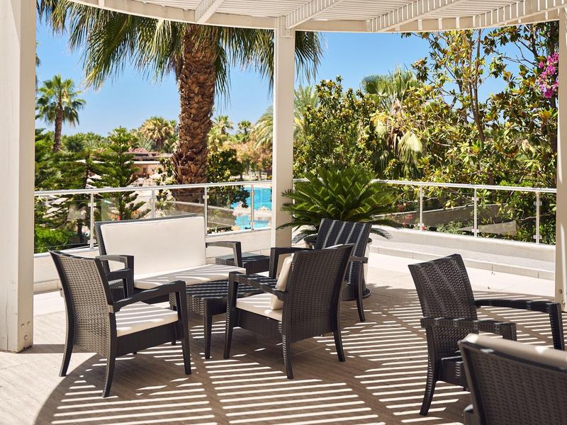 Überdachte Terrasse mit schwarzen Stühlen, weißer Sitzbank und Blick auf Palmen und blauen Himmel.