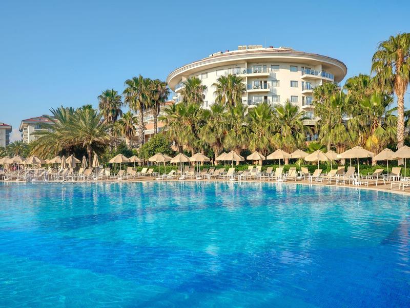 Großer blauer Pool mit Sonnenschirmen, Palmen und rundem Hotelgebäude bei klarem Himmel.