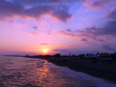 Sonnenuntergang über ruhigem Meer mit heller Sonne, lila Wolken und dunklem Strand im Vordergrund.