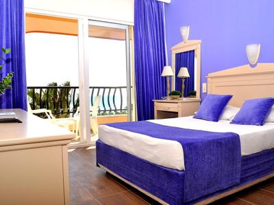 Helles Hotelzimmer mit lila Wänden, Bett und Vorhängen, Balkon mit Meerblick.