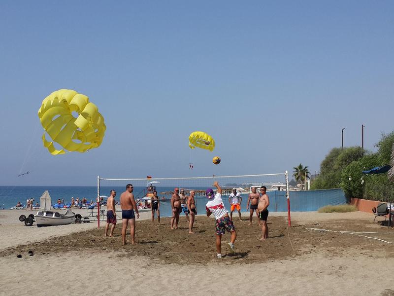 Mehrere Menschen spielen Volleyball am Strand bei klarem Himmel, mit gelben Fallschirmen im Hintergrund.