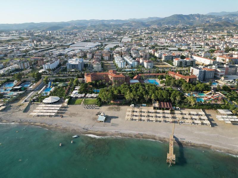 Luftaufnahme eines Hotels mit Pool, Strand und Blick auf eine Küstenstadt und Berge im Hintergrund.