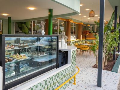 Modernes Café mit gläserner Kuchentheke, grünen Säulen, Pflanzen und weißen Mosaikfliesen.