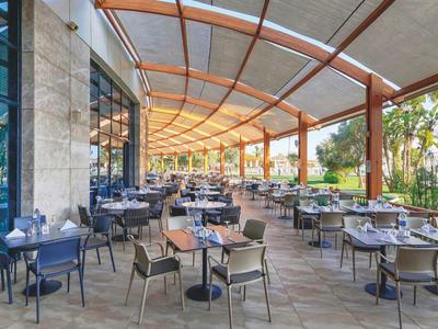 Große Terrasse mit Tischen und Stühlen unter einem gewölbten Holzdach neben Glasfront und Grünflächen.