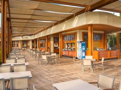 Große, offene Cafeteria mit vielen weißen Tischen und Stühlen, Holzsäulen und Sonnenschutzdach.