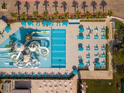 Luftaufnahme eines rechteckigen Pools mit Wasserrutschen, umgeben von blauen Liegestühlen und Palmen.