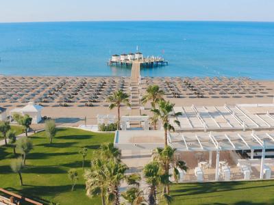 Strand mit Sand, Liegestühlen und Sonnenschirmen, Steg ins Meer, davor Rasen mit Palmen.