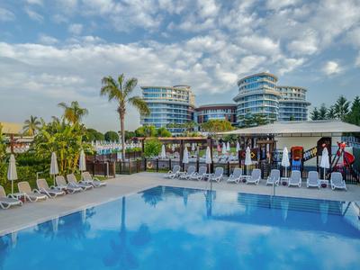 Moderner Hotelpoolbereich mit Liegestühlen, Palmen und zwei hohen Gebäuden im Hintergrund.