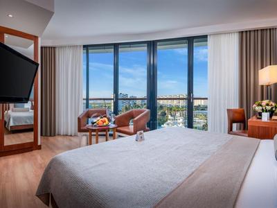 Helles Hotelzimmer mit Doppelbett, Sitzbereich und Fenster mit Blick auf Stadt und Himmel.