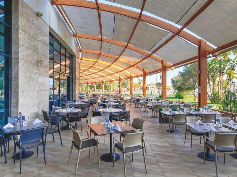 Große Terrasse mit Tischen und Stühlen unter einem gewölbten Holzdach neben Glasfront und Grünflächen.