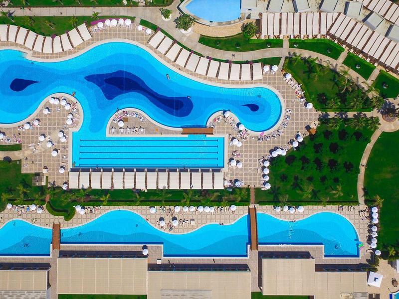 Luftaufnahme eines Pools mit geschwungenen und rechteckigen Becken, umgeben von Liegestühlen und Grünflächen.