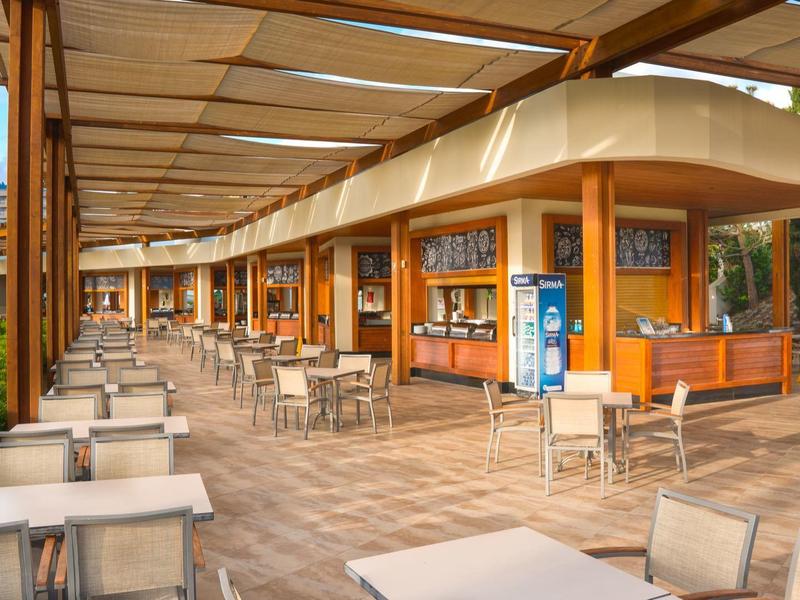 Große, offene Cafeteria mit vielen weißen Tischen und Stühlen, Holzsäulen und Sonnenschutzdach.