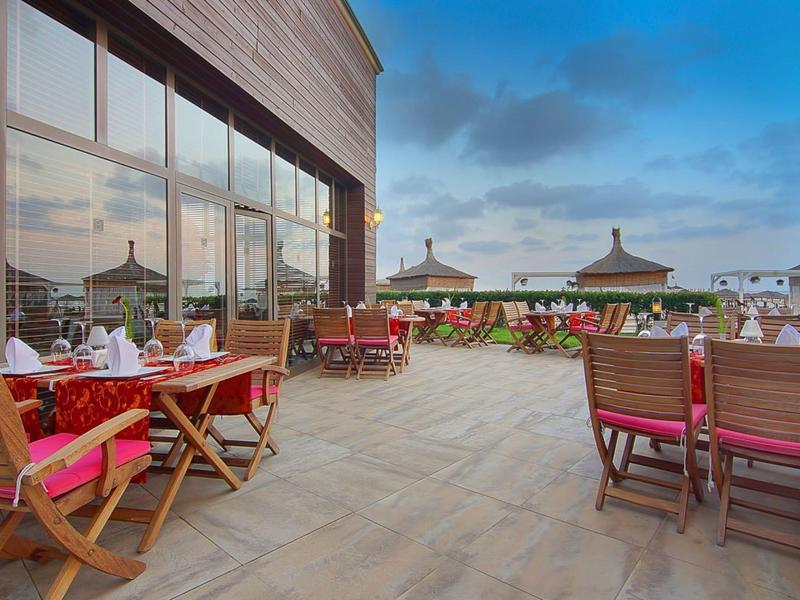 Terrasse mit Holztischen und roten Kissen, Glasfassade, blauer Himmel und Backsteinmauer mit Pflanzen.