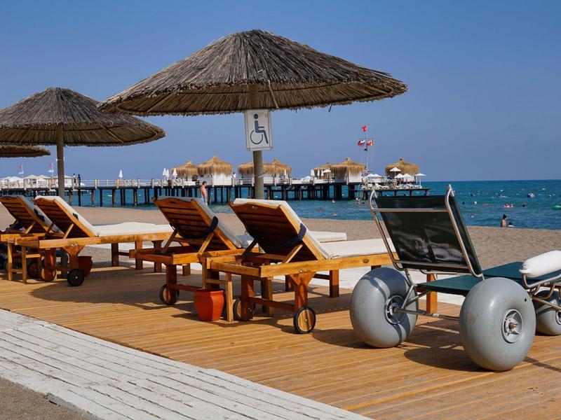 Strandliegen mit Strohdächern und ein Strandrollstuhl auf Holzsteg am Meer bei klarem Himmel.