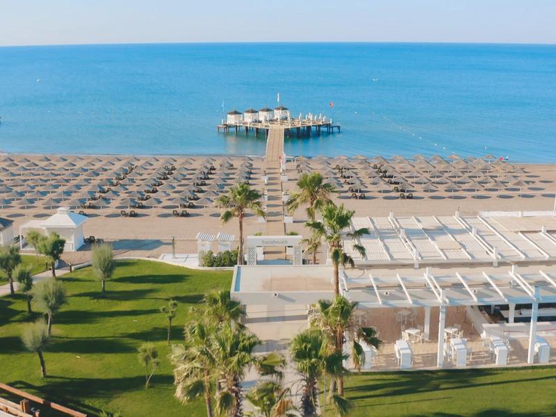 Strand mit Sand, Liegestühlen und Sonnenschirmen, Steg ins Meer, davor Rasen mit Palmen.