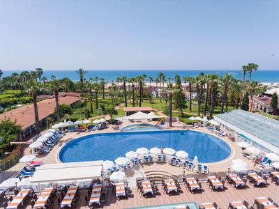 Großer Pool mit weißen Sonnenschirmen und Liegen, umgeben von Palmen und Meer im Hintergrund.