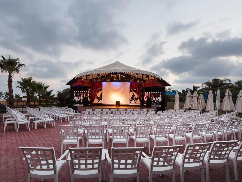 Freiluftbühne mit zahlreichen weißen Stühlen und geschlossenen Sonnenschirmen unter bewölktem Himmel.